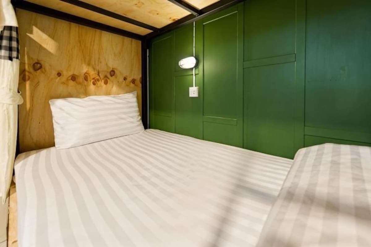 Una cama individual con colcha a rayas, cabecero de madera y pared verde con lámpara.
