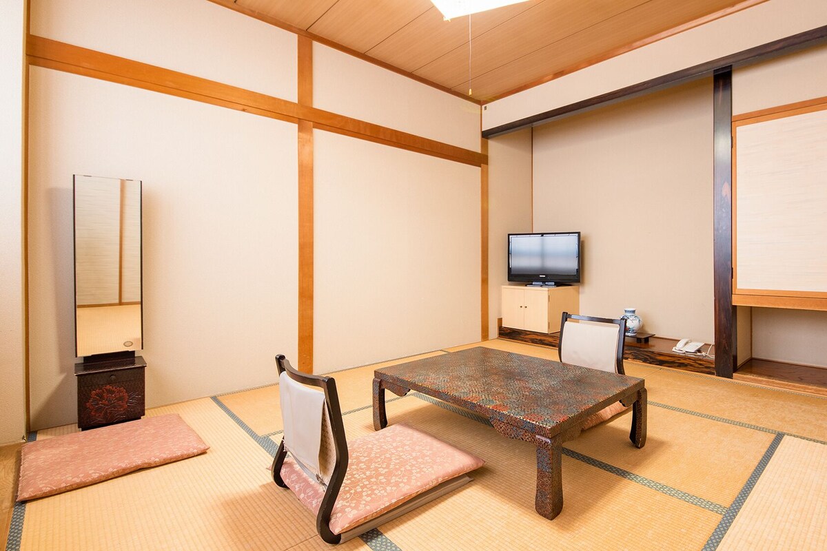 A traditional Japanese room with tatami flooring, a low table, a chair, a television, and a cabinet.