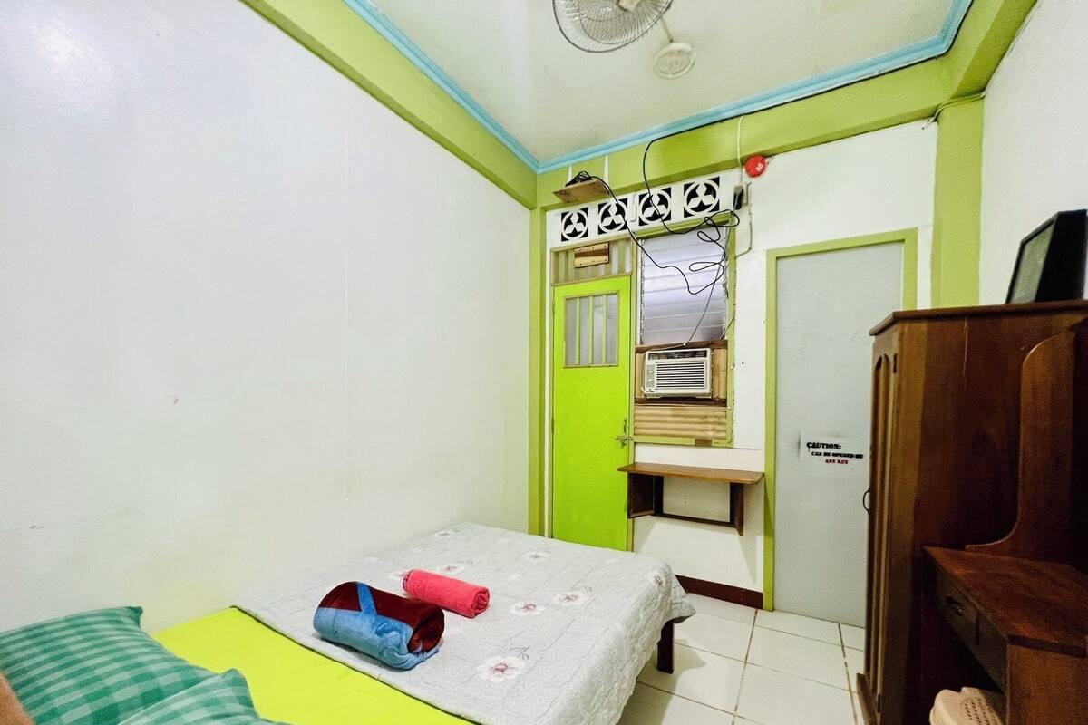 Una habitación pequeña y sencilla con una cama, un ventilador y un armario de madera.