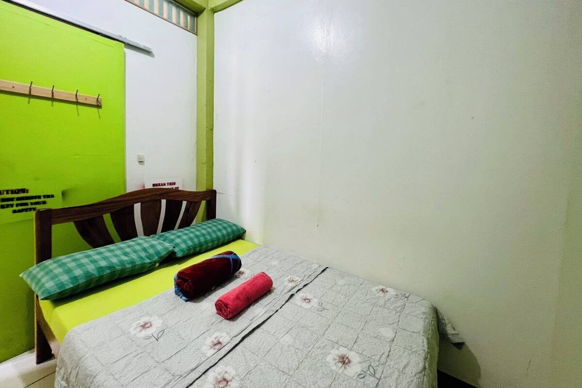 Una habitación pequeña con una cama individual, puerta verde, cabecero de madera y colcha con estampado floral.