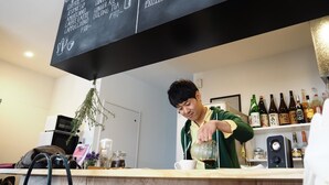 A bar area with a chalkboard menu listing coffee and alcohol options, a person pouring a drink, and a shelf with bottles of alcohol.
