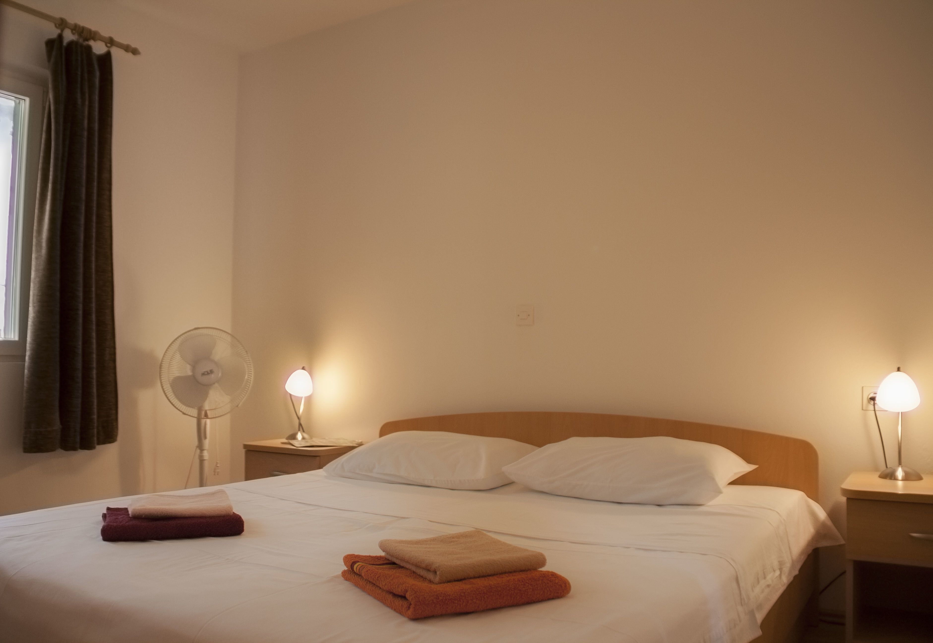 A neatly made bed with white and brown towels, a bedside fan, and a lamp on a wooden nightstand.