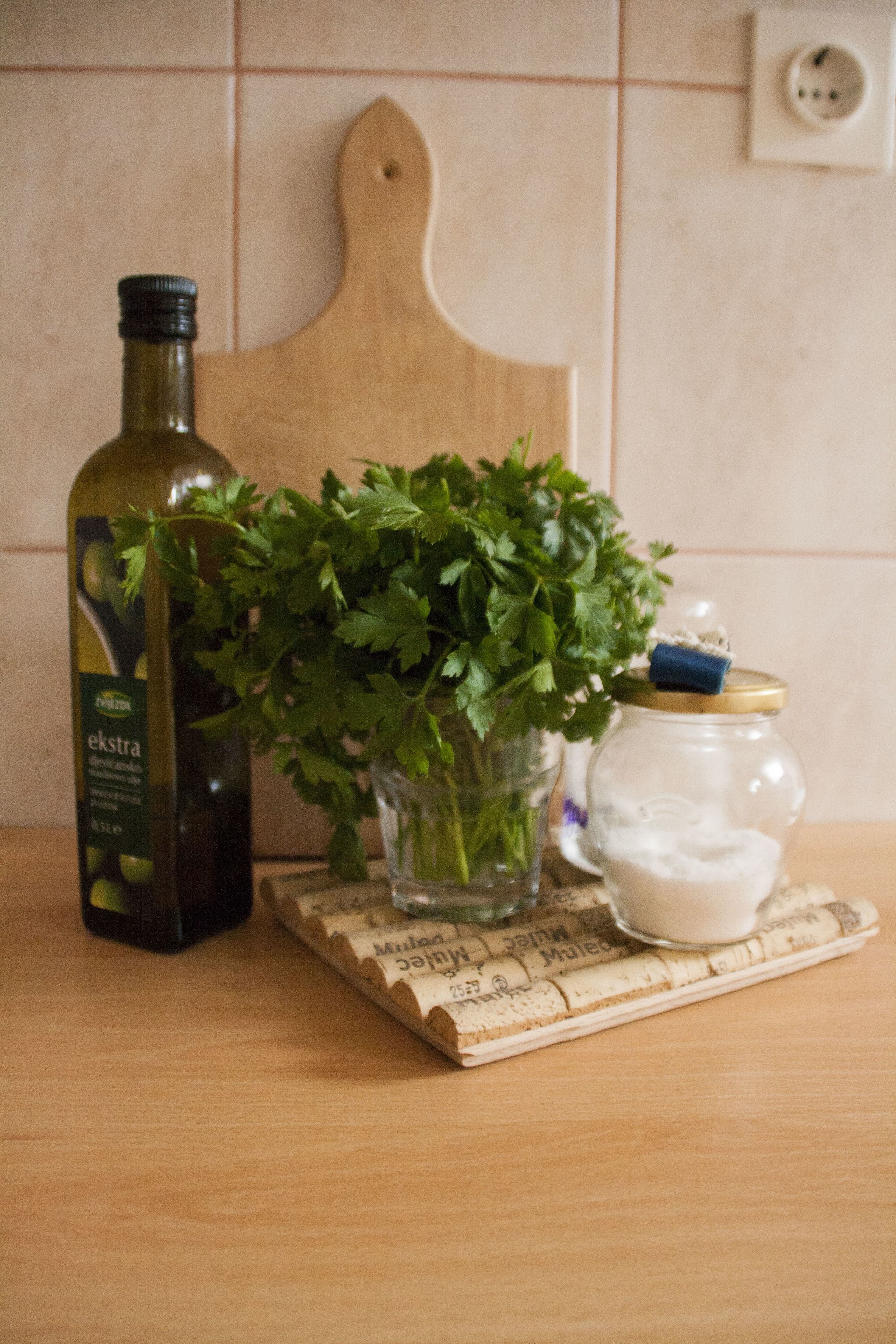 Eine Flasche Olivenöl, ein Glas Petersilie und ein Glas weiße Sahne auf einer Korktafel.