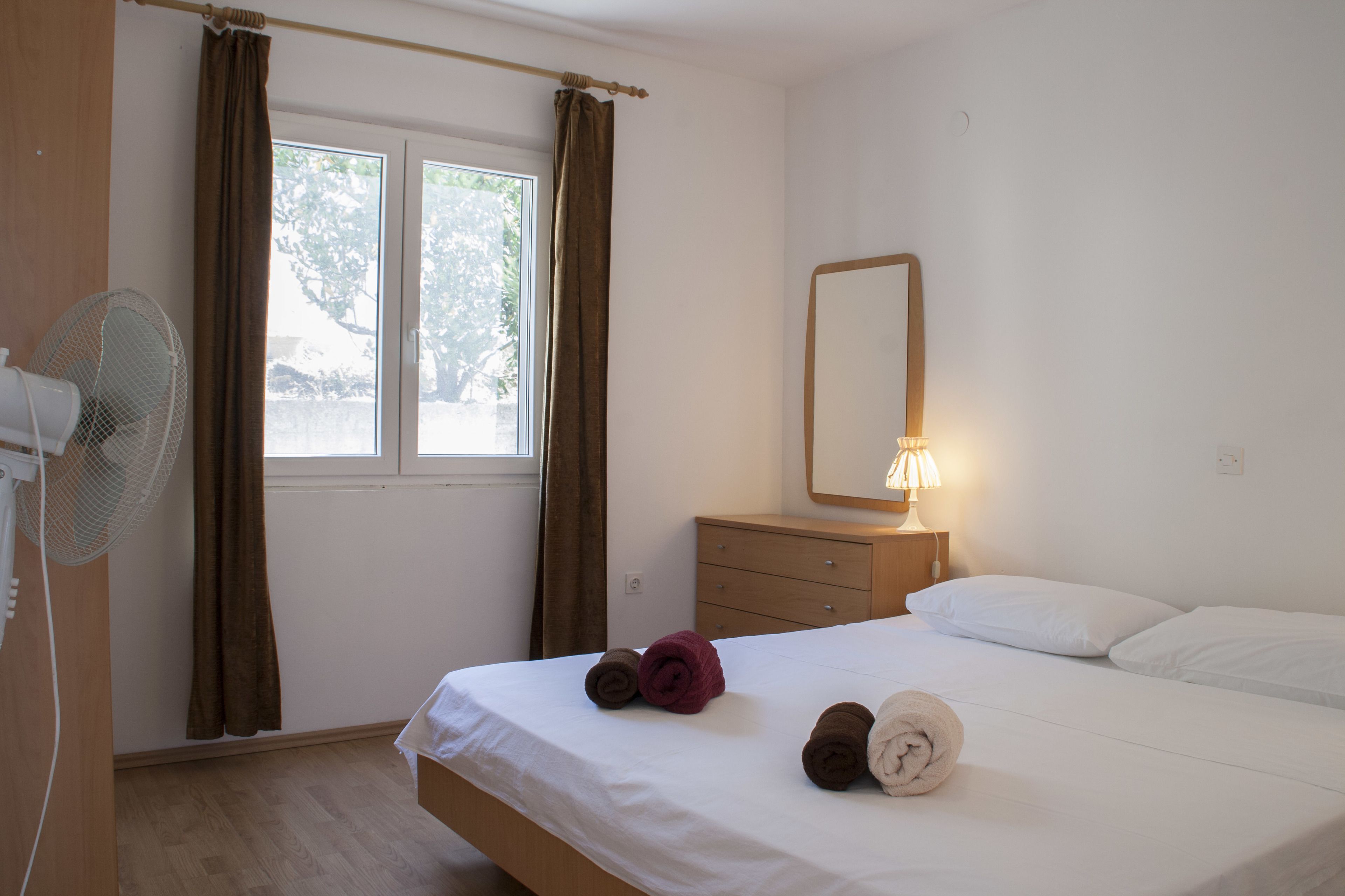 Une chambre d’hôtel avec un lit, une table de chevet avec une lampe, un ventilateur et une fenêtre avec des rideaux.
