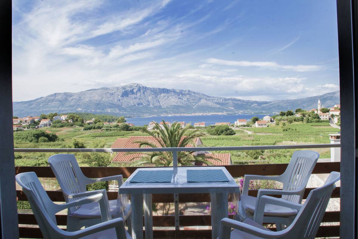 En balkon med to hvide plastikstole og et bord, med udsigt over et bjerglandskab og et vandområde.