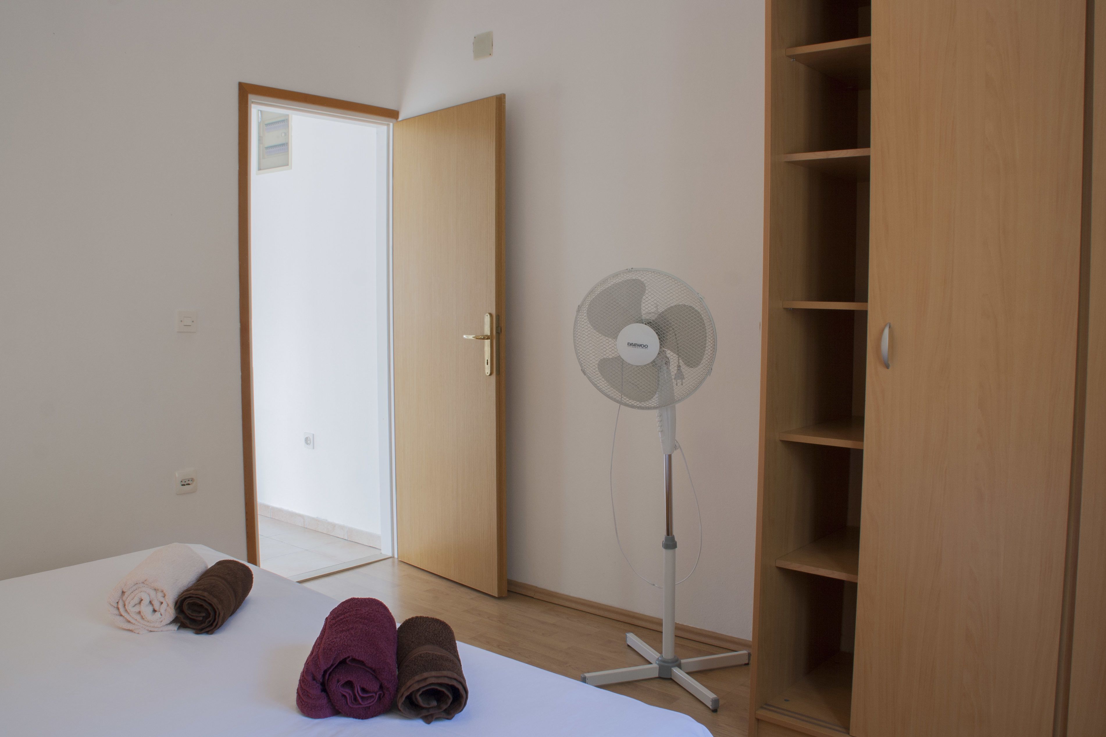 A hotel room with a bed, rolled towels, a standing fan, and a wooden wardrobe.