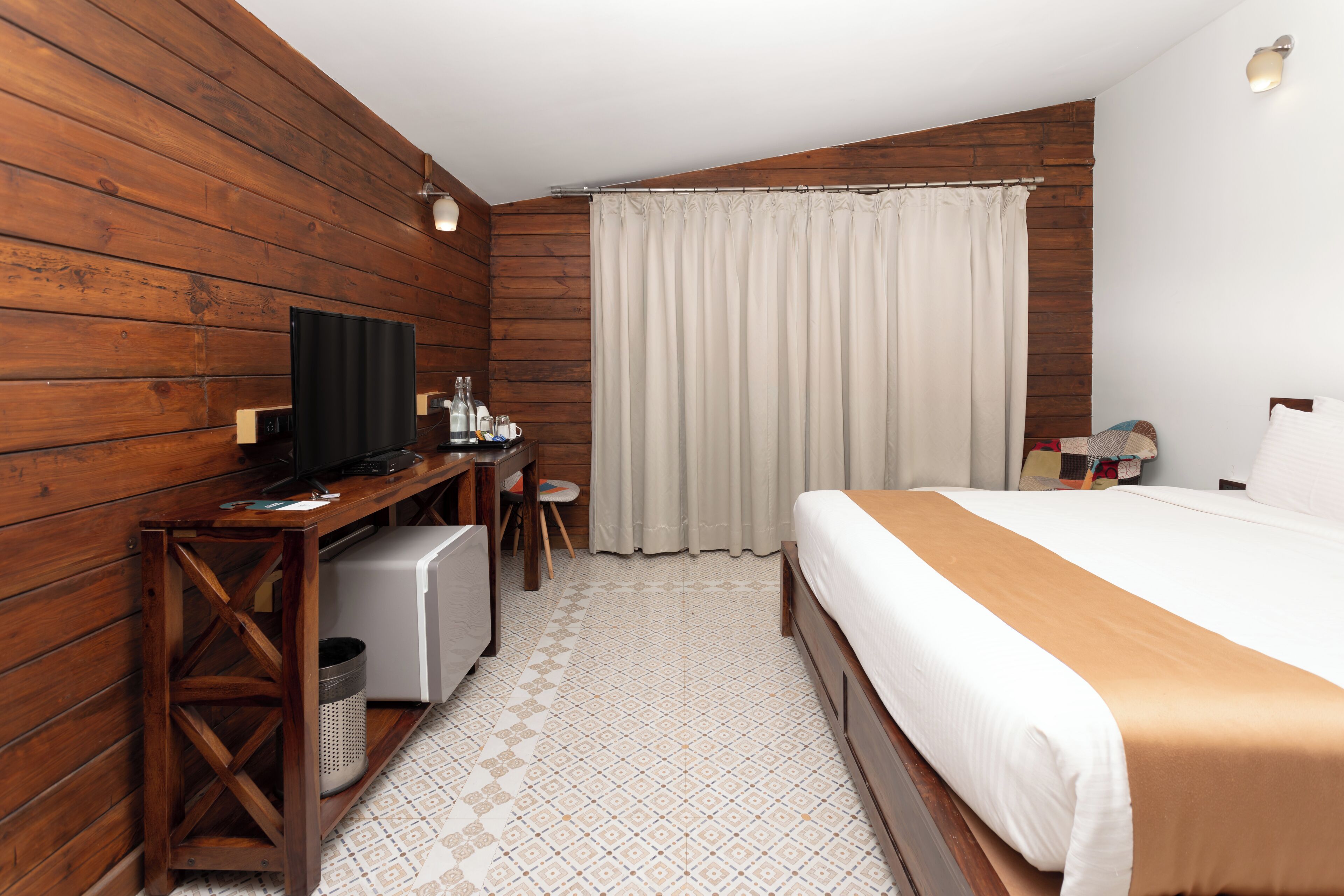 A room with wooden walls, a bed with a white and brown bedspread, a desk with a television, and a window with curtains.