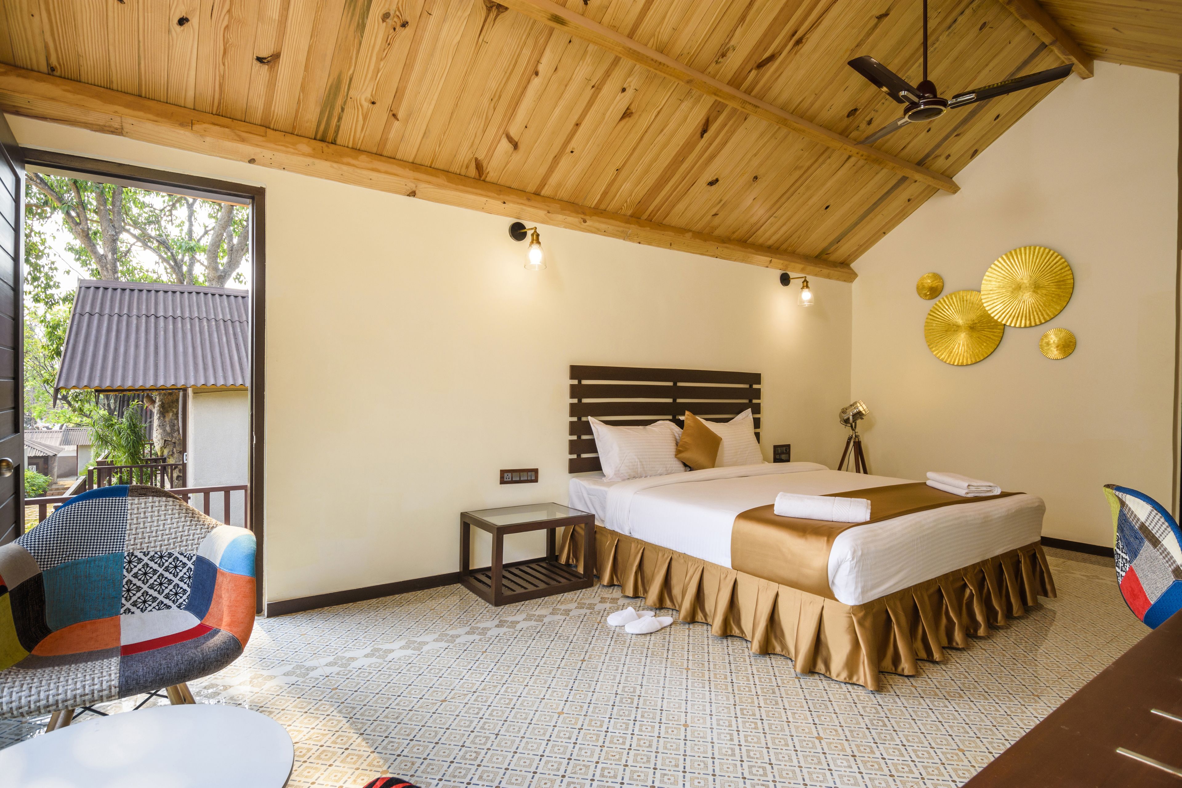A bedroom with a wooden ceiling, a bed with a beige bedspread, a nightstand, a chair, and a window with a view of trees.
