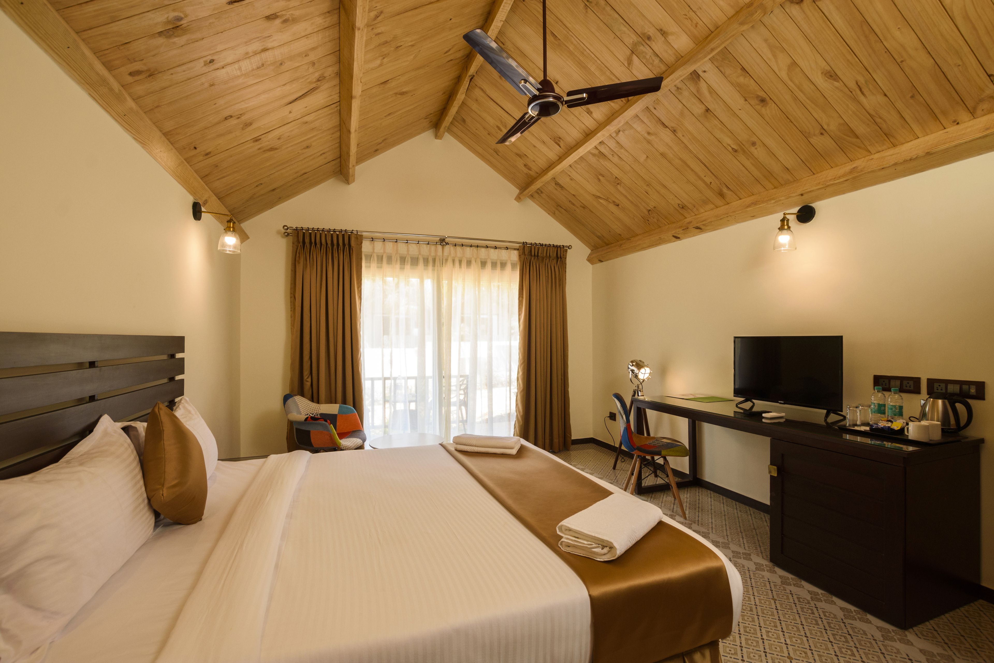 Une chambre d’hôtel avec un plafond en bois, un lit avec du linge de maison blanc, un bureau avec une télévision et une fenêtre avec des rideaux.