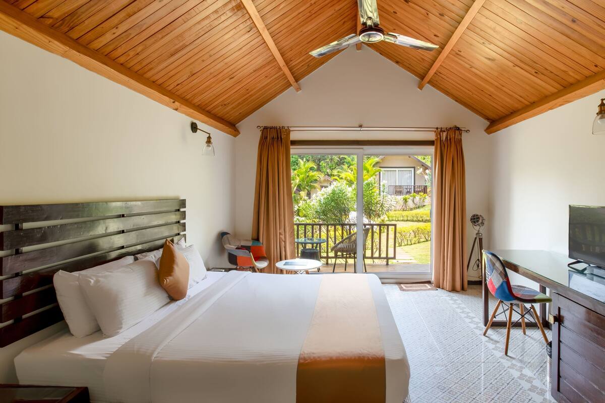 A hotel room with a wooden ceiling, a bed with white bedding, a desk with a chair, and a balcony with a view of greenery.