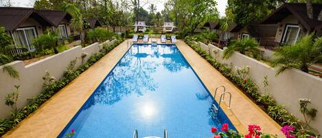 A rectangular swimming pool with a ladder leading into it, surrounded by lush greenery and small buildings with balconies.