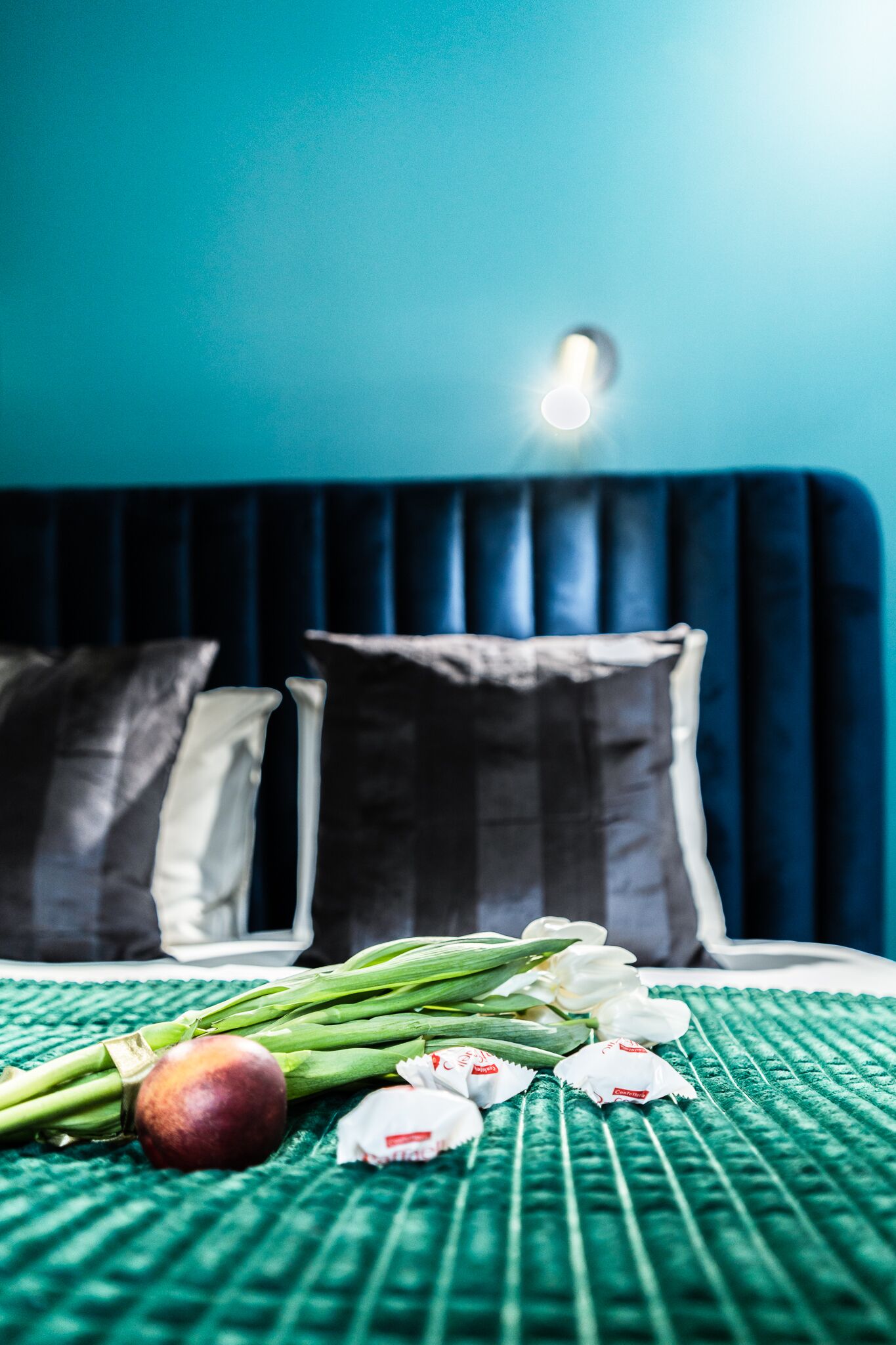 A bed with a green textured bedspread, a bouquet of white flowers, and several wrapped chocolates on the bed.