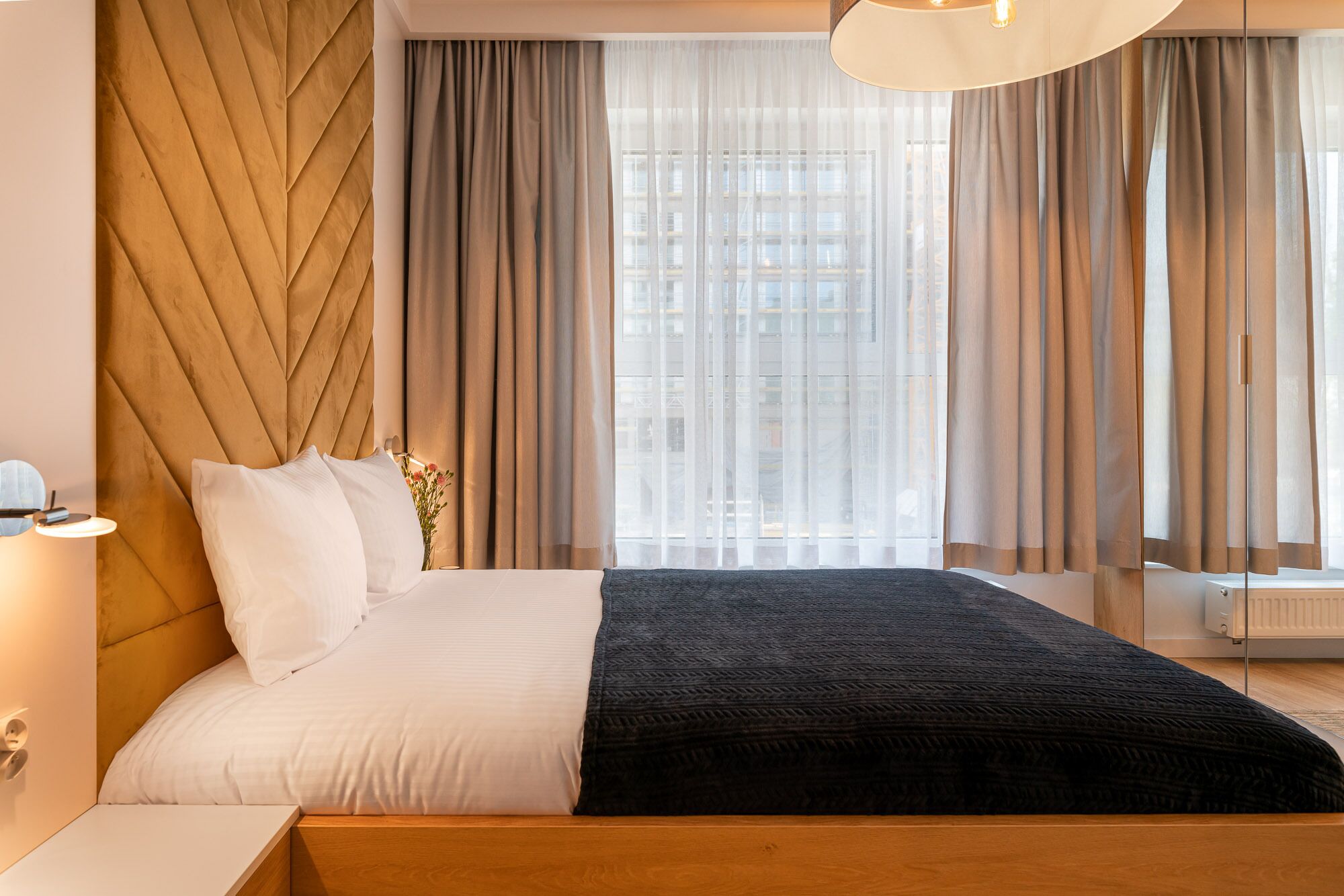 A modern hotel room with a wooden headboard, a large bed with white and black bedding, a bedside table with a lamp, and sheer curtains allowing natural light.