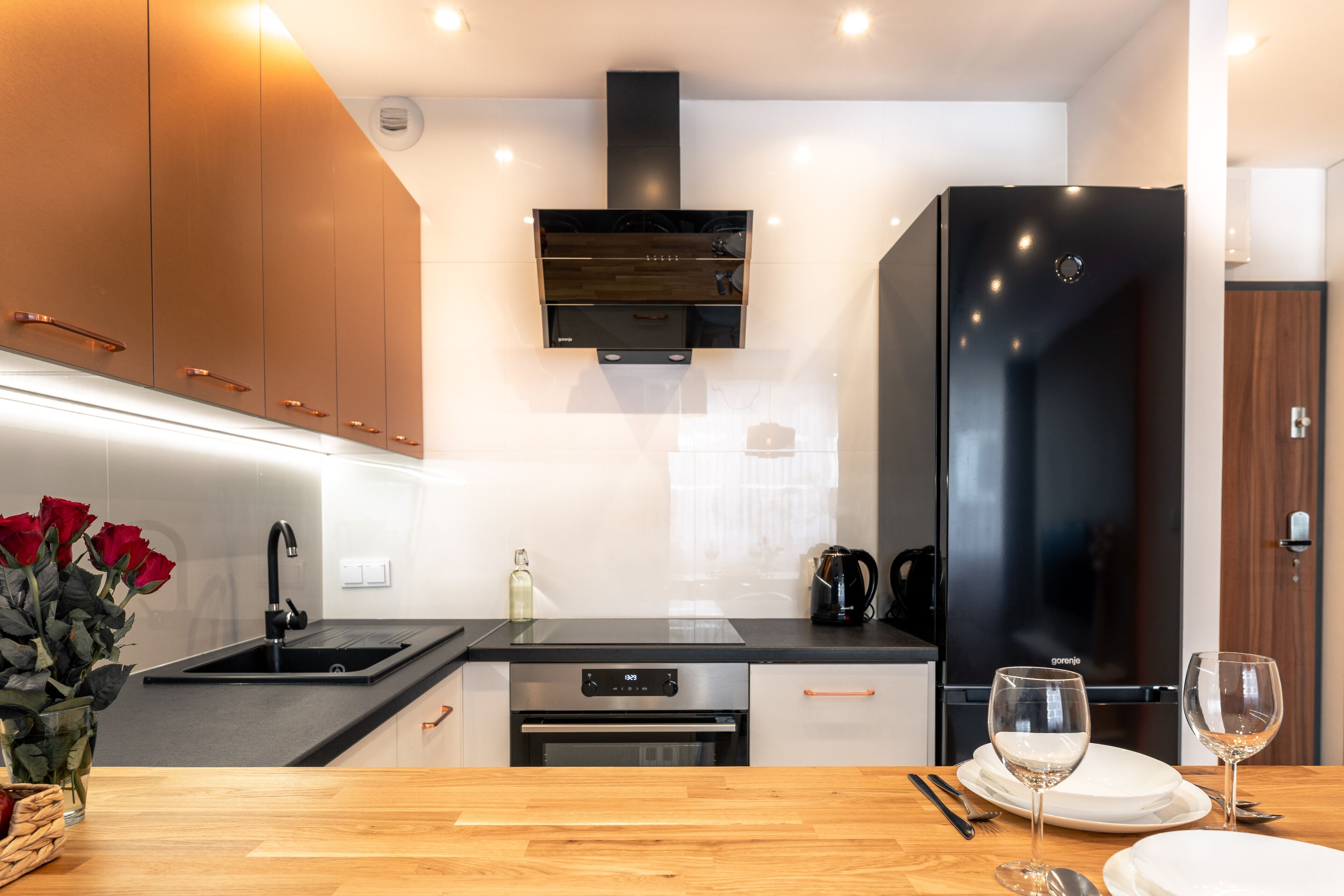 Una cocina moderna con armarios de madera, un refrigerador negro y un comedor con una mesa de madera y copas de vino.