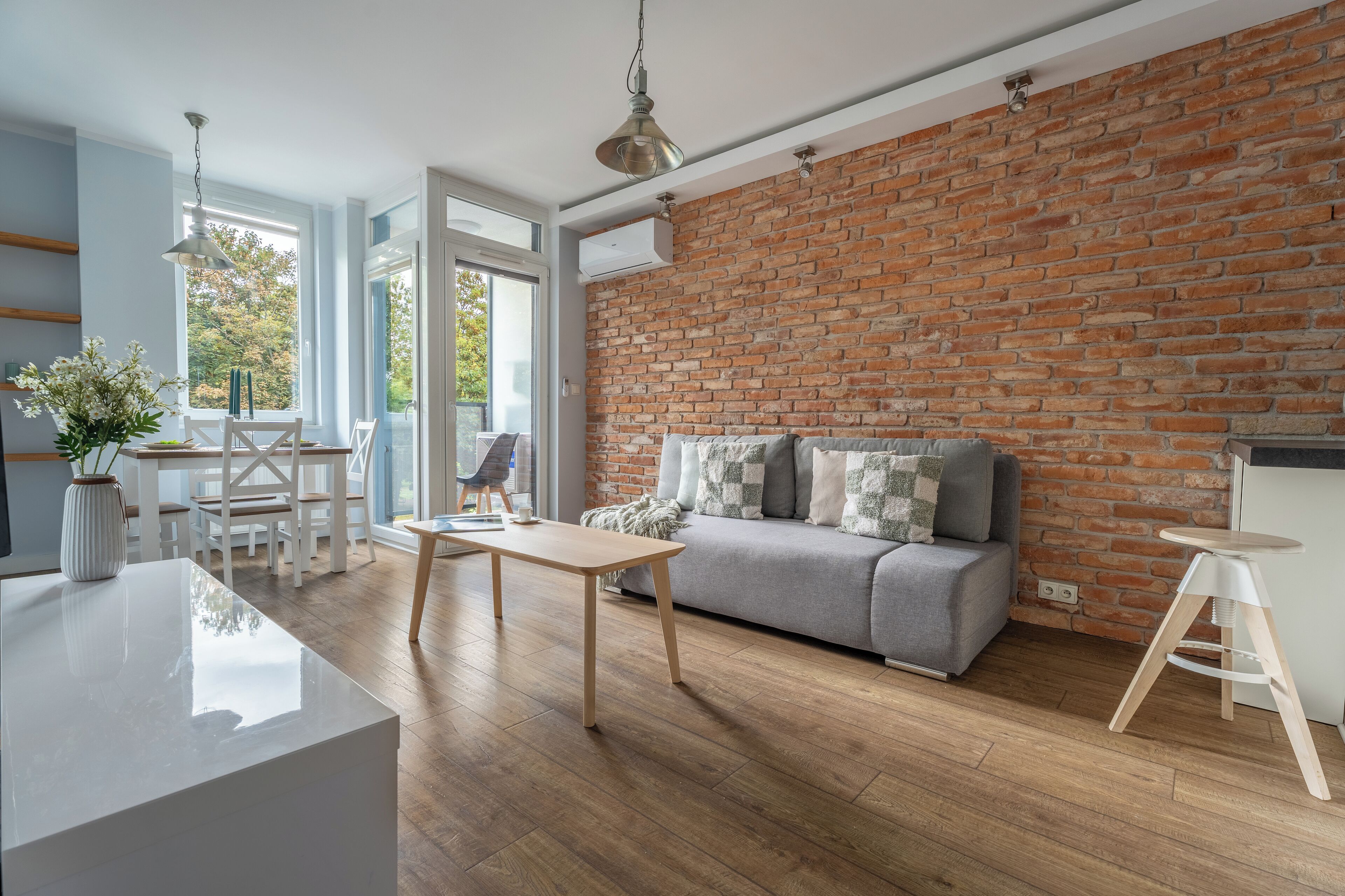 Una sala de estar moderna con una pared de ladrillo, un sofá gris, piso de madera y una mesa blanca.