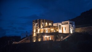 A modern building with stone walls and multiple windows, situated on a hillside. - Tinos Pegion House (Tinos)