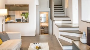 A modern living room with a white sofa, a coffee table with decorative items, a staircase leading to an upper level, and a kitchen area visible through an open doorway. - Tinos Pegion House (Tinos)