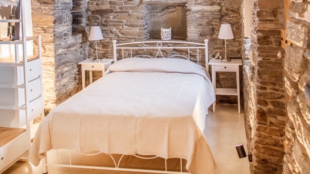 A bedroom with stone walls, a bed, bedside tables, and a window with a decorative arch. - Tinos Pegion House (Tinos)