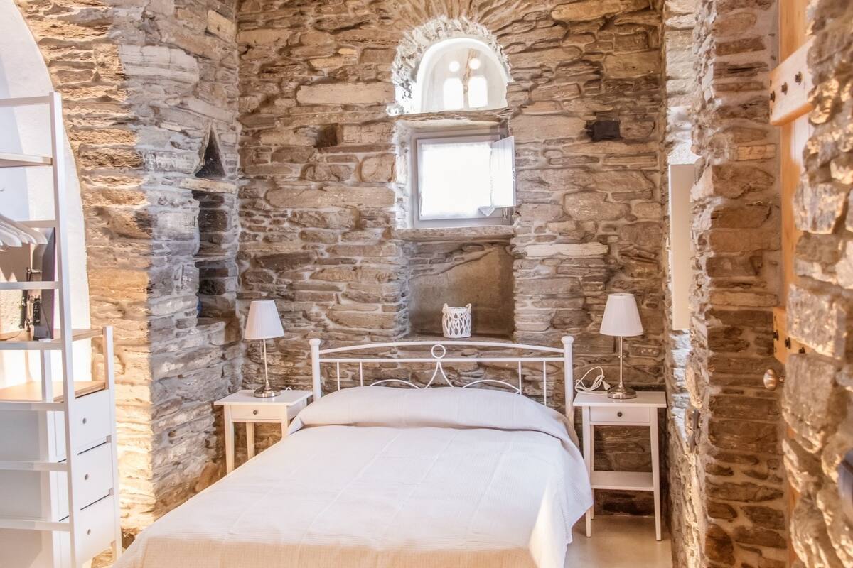 Une chambre avec des murs en pierre, un lit, des tables de chevet et une fenêtre surmontée d’une arche décorative.