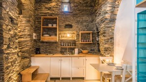 A cozy kitchen with stone walls, wooden shelves, and a small dining area with a table and benches. - Tinos Pegion House (Tinos)