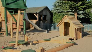 A wooden play structure with a slide and climbing elements, a small wooden house, and a fenced area. - Das Fritz Hotel (Schwerin)
