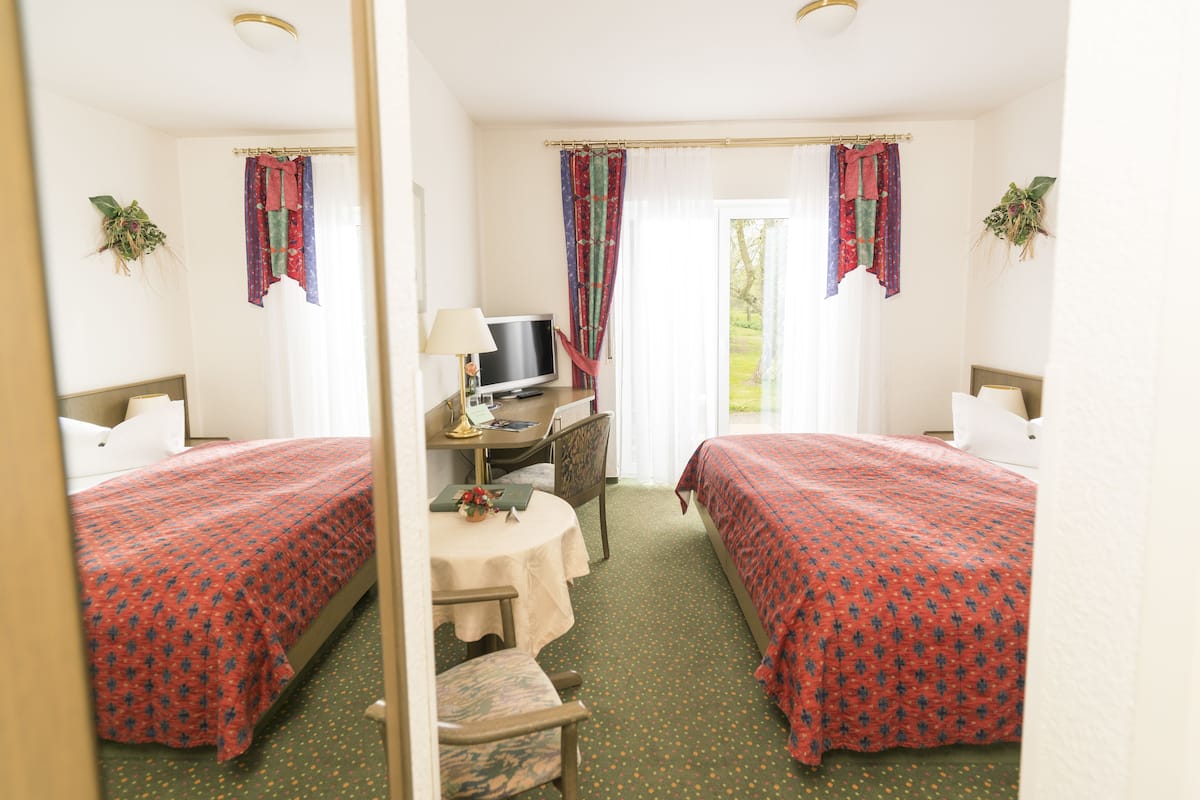 A hotel room with two beds, a desk with a TV, a chair, and a small table with a lamp.