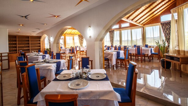 A spacious dining area with multiple tables and chairs, large windows with curtains, and a ceiling with star-shaped light fixtures. - Hotel Europa Kokeltal (Albesti)