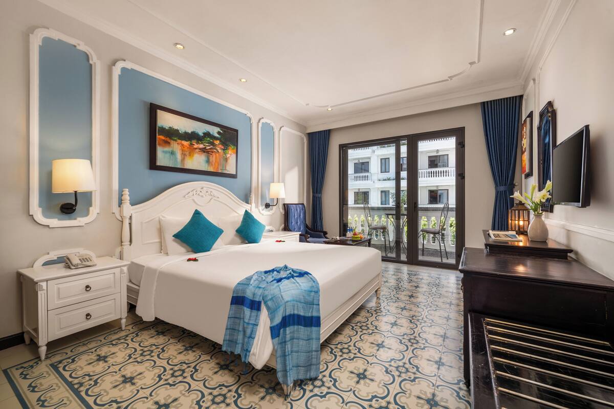 A hotel room with a large bed, a nightstand with a phone, a desk with a lamp, and a view of the outside balcony.