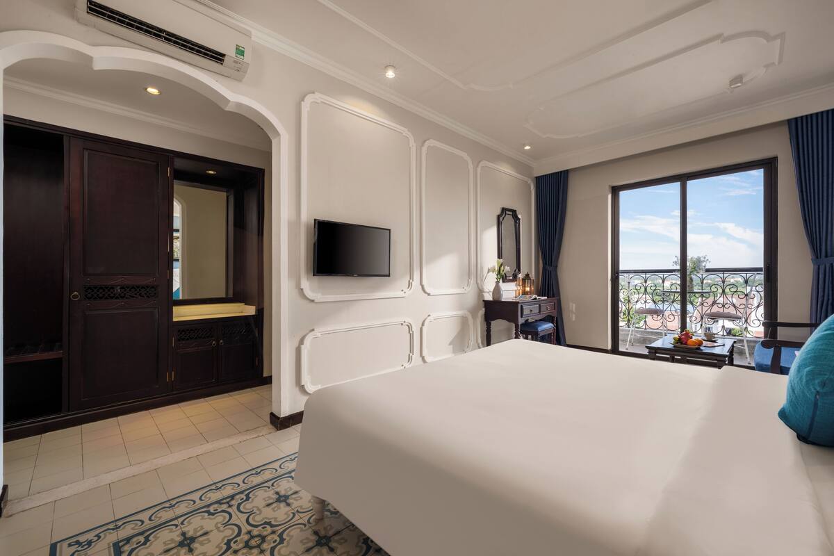 A hotel room with a large bed, a television, a desk with a chair, a window with a balcony view, and a decorative mirror.