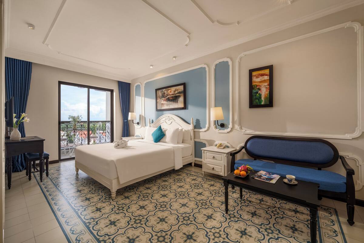 A hotel room with a bed, a bedside table, a chair, a sofa, a balcony, and a view of the outdoors.