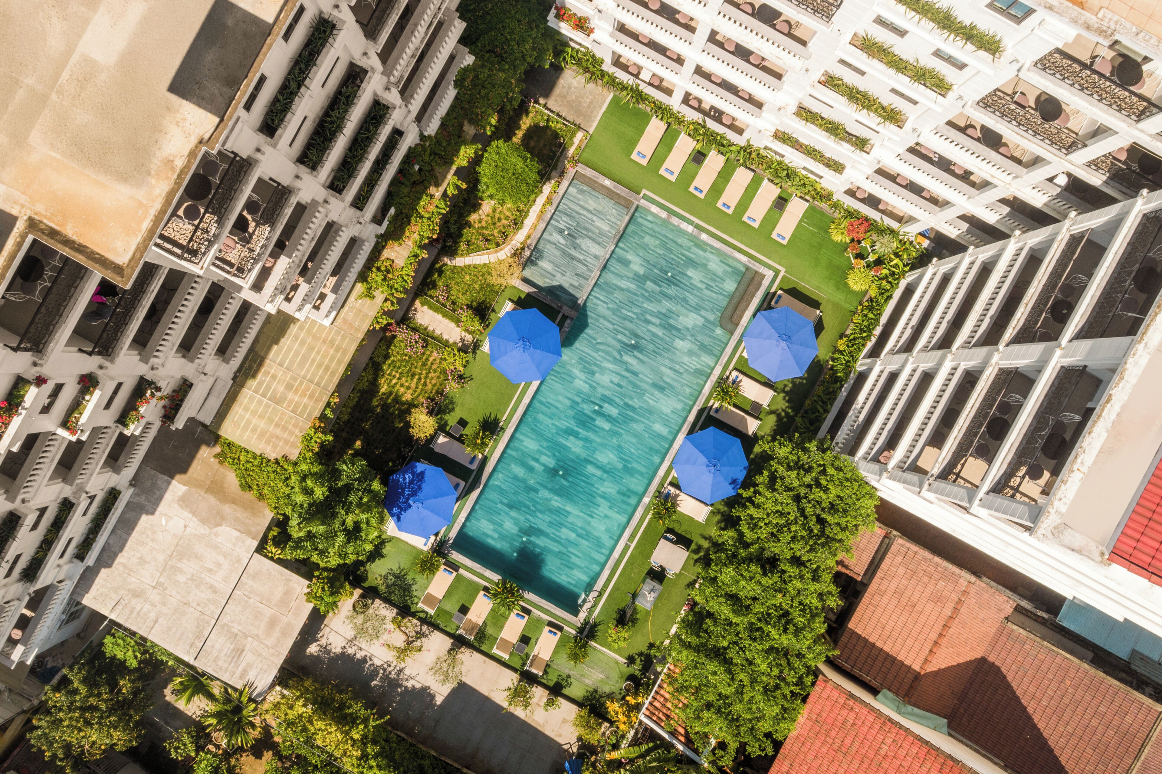 An aerial view of a hotel with a central swimming pool, surrounded by lush greenery and multiple balconies.