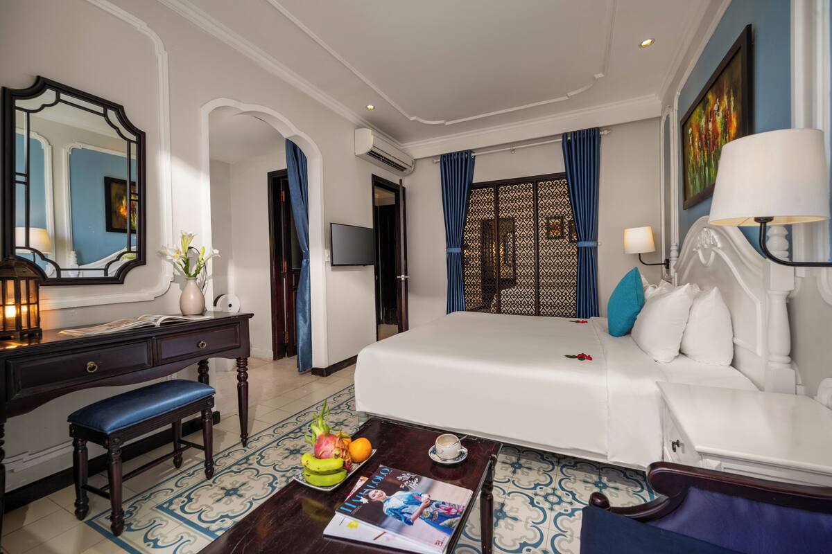 A hotel room with a large bed, a desk with a chair, a small table with a magazine, and a decorative mirror.