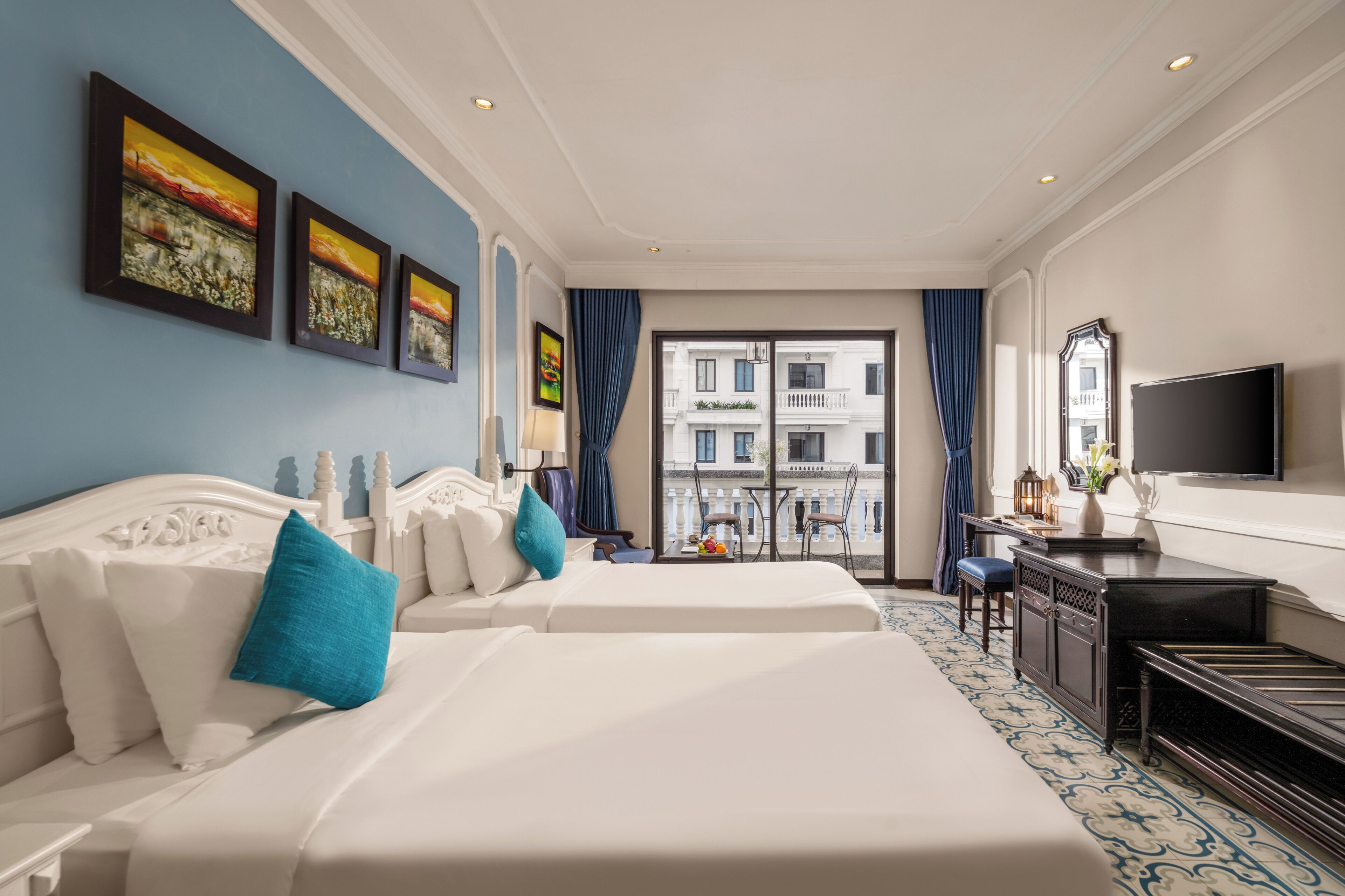 Ein Hotelzimmer mit zwei Betten, einem Schreibtisch, einem Fernseher und einem Balkon mit Blick auf Gebäude.