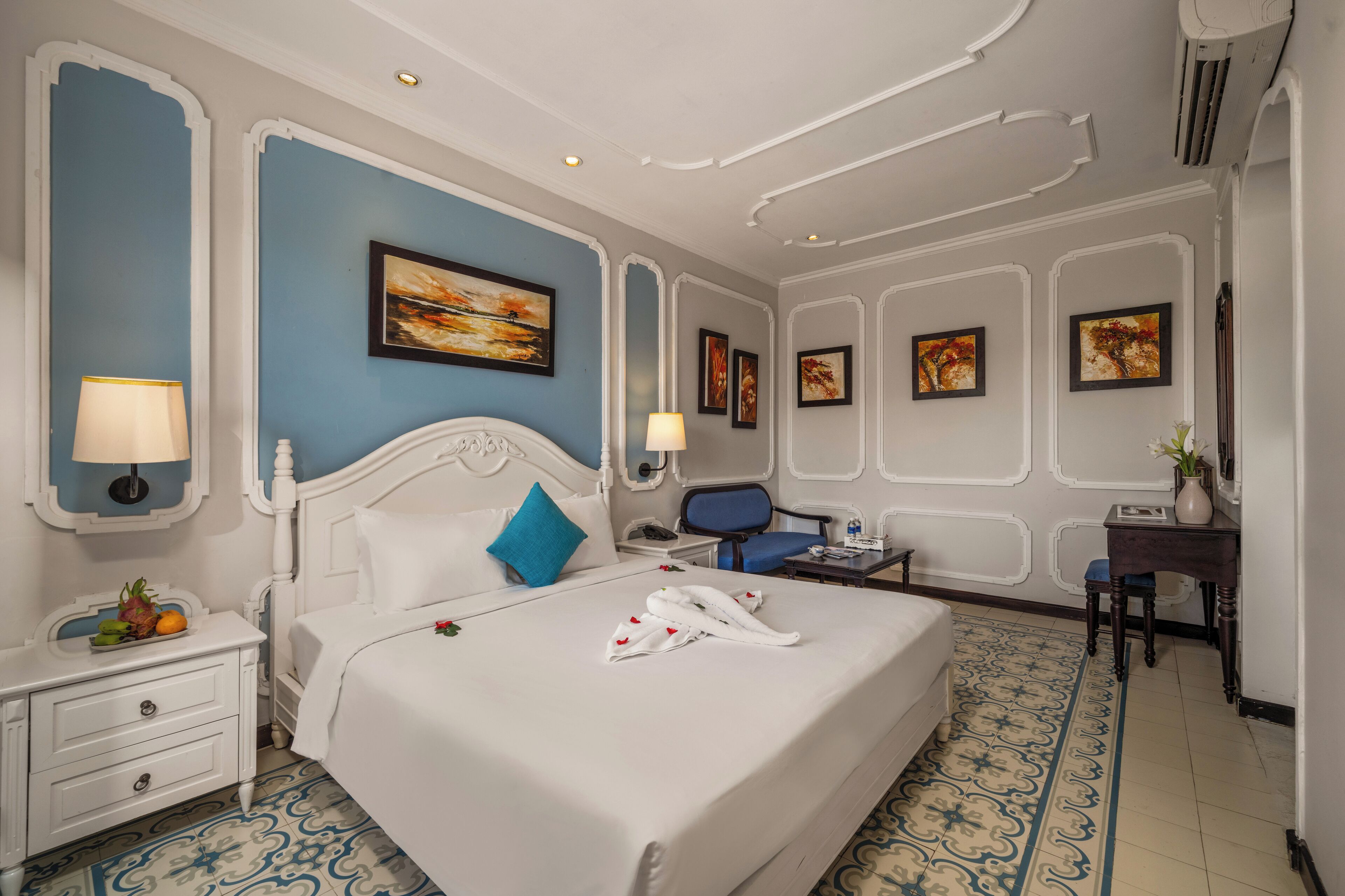 Une chambre d’hôtel avec un grand lit, deux tables de chevet, un bureau et une chaise. Les murs sont ornés de tableaux encadrés et un vase rempli de fleurs est posé sur le bureau.