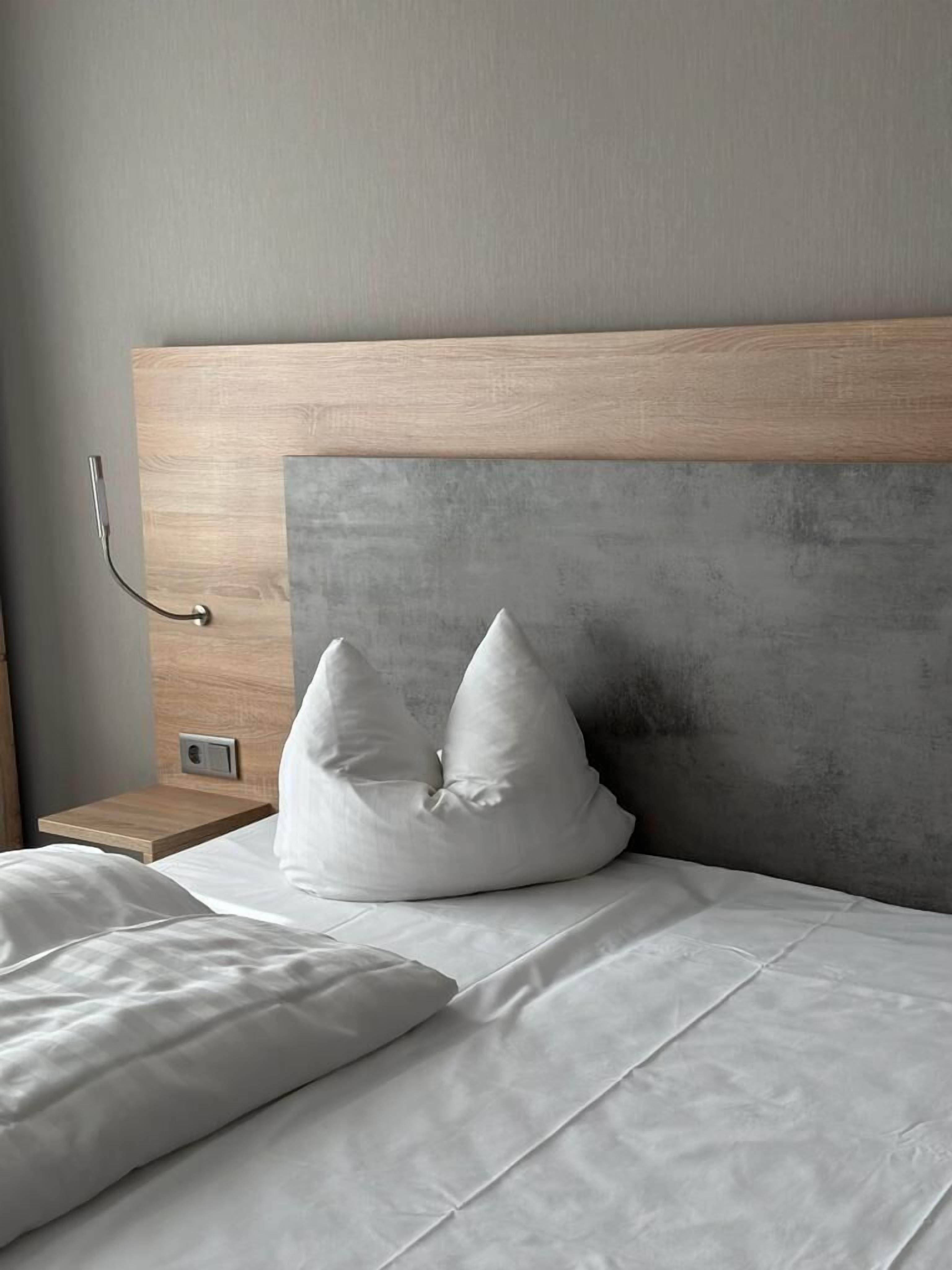 A neatly made bed with white linens and two white pillows, a wooden headboard, and a small bedside table with a power outlet.