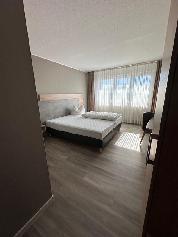 A modern hotel room with a large bed, a wooden headboard, a bedside table, and a view of the outdoors through sheer curtains. - Rheinhessen Inn (Woerrstadt)
