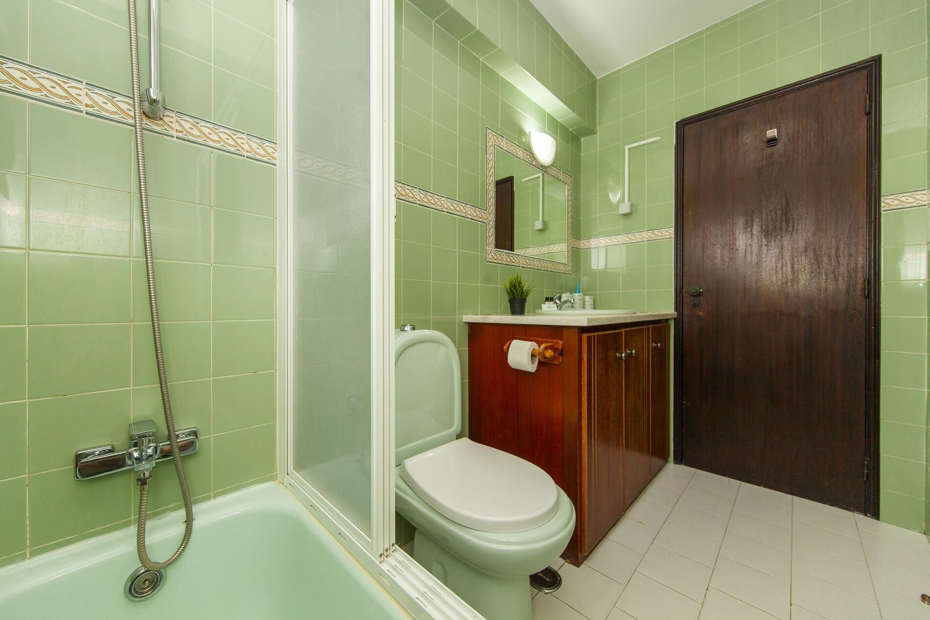 A bathroom with green tiles, a white toilet, a wooden cabinet, and a shower with a hose.