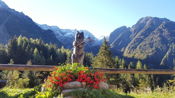 Ein hölzerner Zaun mit einer Statue eines Bären, der eine Axt hält, umgeben von roten Blumen und Grünpflanzen, mit einer Berglandschaft im Hintergrund.