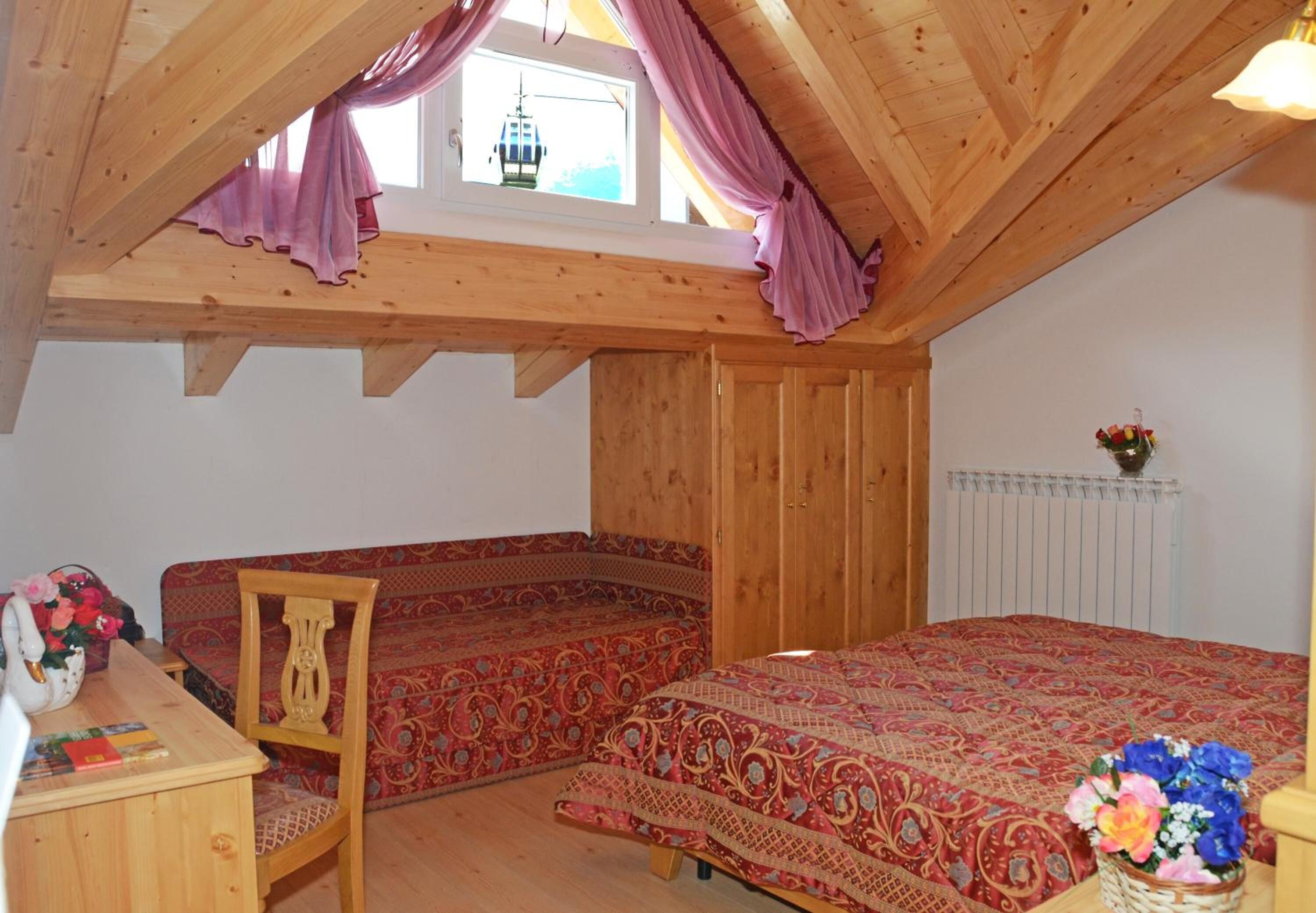 Ein gemütliches Schlafzimmer im Dachgeschoss mit Holzbalkendecke, einem roten, gemusterten Bett, einem Holzstuhl und einem kleinen Tisch mit Blumenvase.