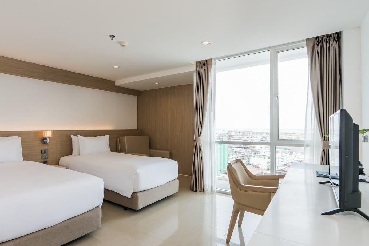 A hotel room with two beds, a desk with a TV, and a large window offering a city view.