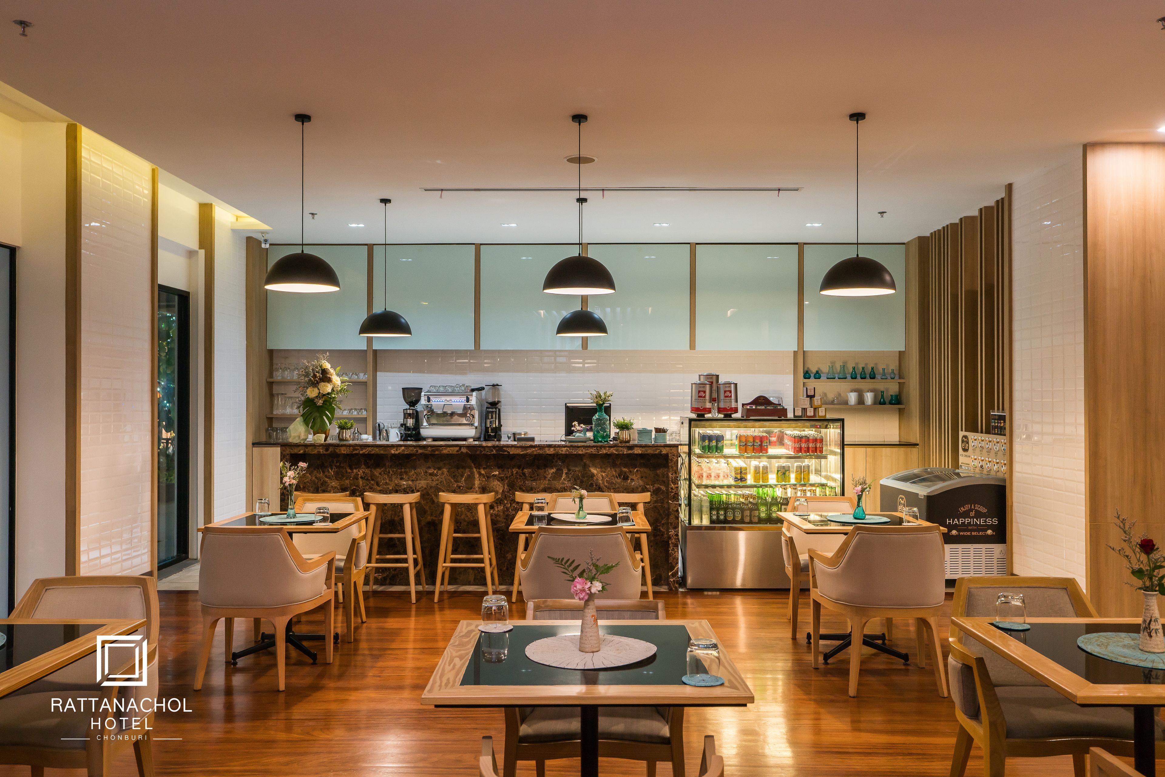 Un restaurante moderno de hotel con mobiliario de madera, iluminación colgante y una zona de bar con mostrador refrigerado.