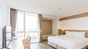 A hotel room with a bed, a desk, a chair, a TV, and a large window with curtains. - Rattanachol Hotel (Chonburi)