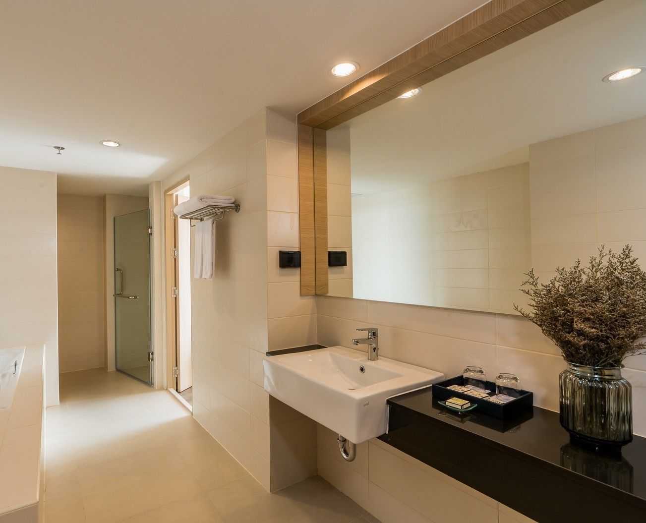A modern bathroom with a white sink, a large mirror, and a vase with dried flowers.