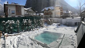 Zona exterior cubierta de nieve con un jacuzzi, rodeada de edificios y montañas.