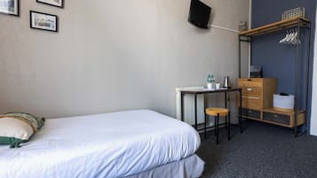 Une chambre d’hôtel bien rangée, avec un lit, un bureau, une chaise et une armoire où sont suspendus des vêtements.