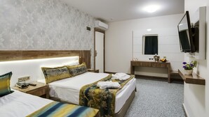 Una habitación de hotel con dos camas, cabecera de madera, mesita de noche con un libro y un control remoto, tocador con espejo y una planta pequeña.