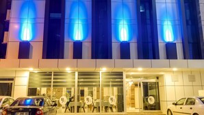 Un edificio moderno con luces azules iluminadas en la fachada, un área de estacionamiento con coches, y una entrada visible con puertas de cristal.