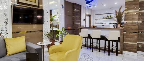 A modern hotel lobby with a bar area, a sofa, a yellow armchair, and a TV mounted on the wall.