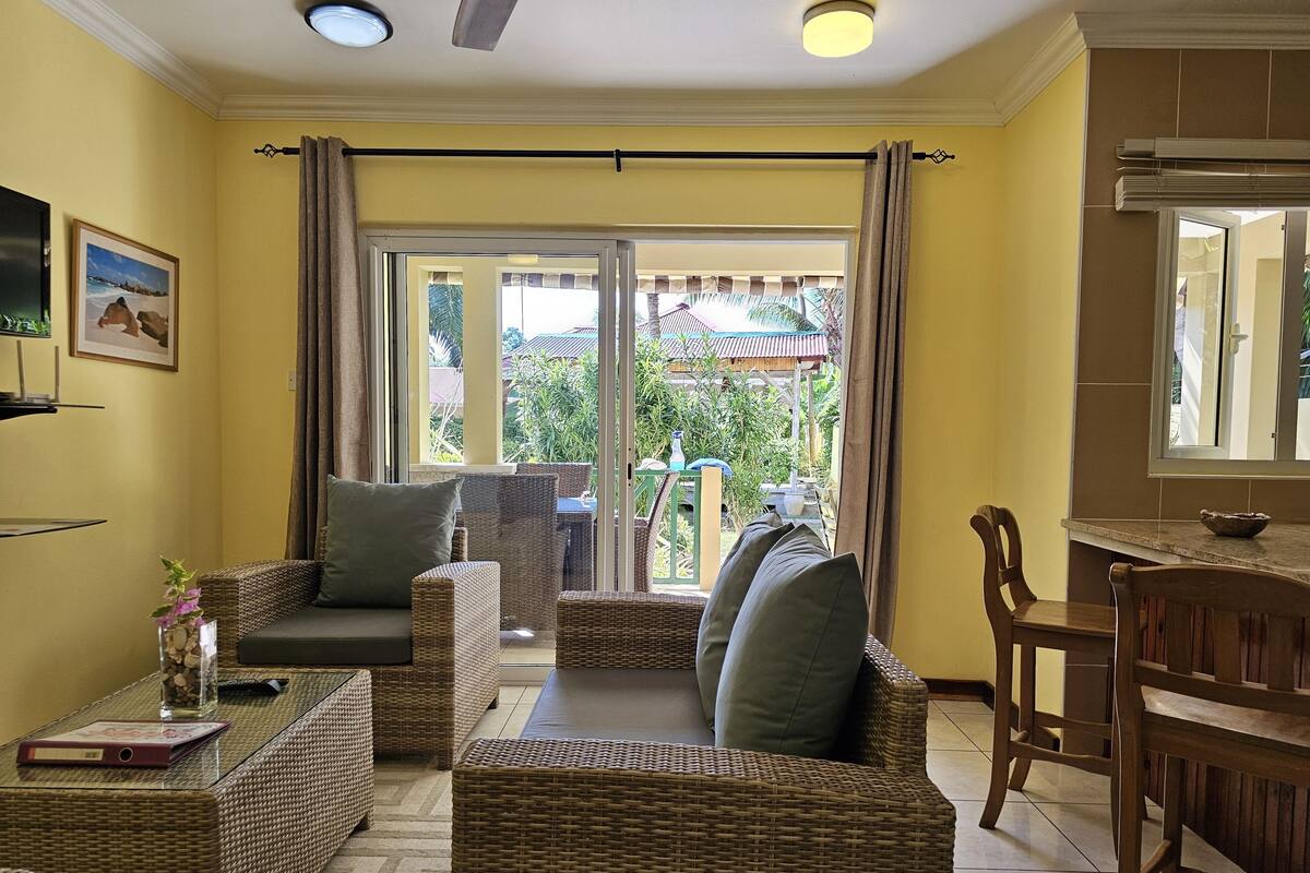 A living room with wicker furniture, a glass-top coffee table, and a dining area with a wooden table and chairs.