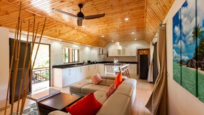 Una cocina moderna con armarios blancos, un comedor con mesa y sillas blancas, y una sala de estar con sofá color beige y cojines rojos.